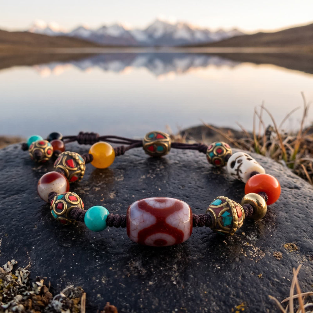 Three-eyed red dzi cord bracelet on a dark stone at the edge of a still mountain lake reflecting snow peaks