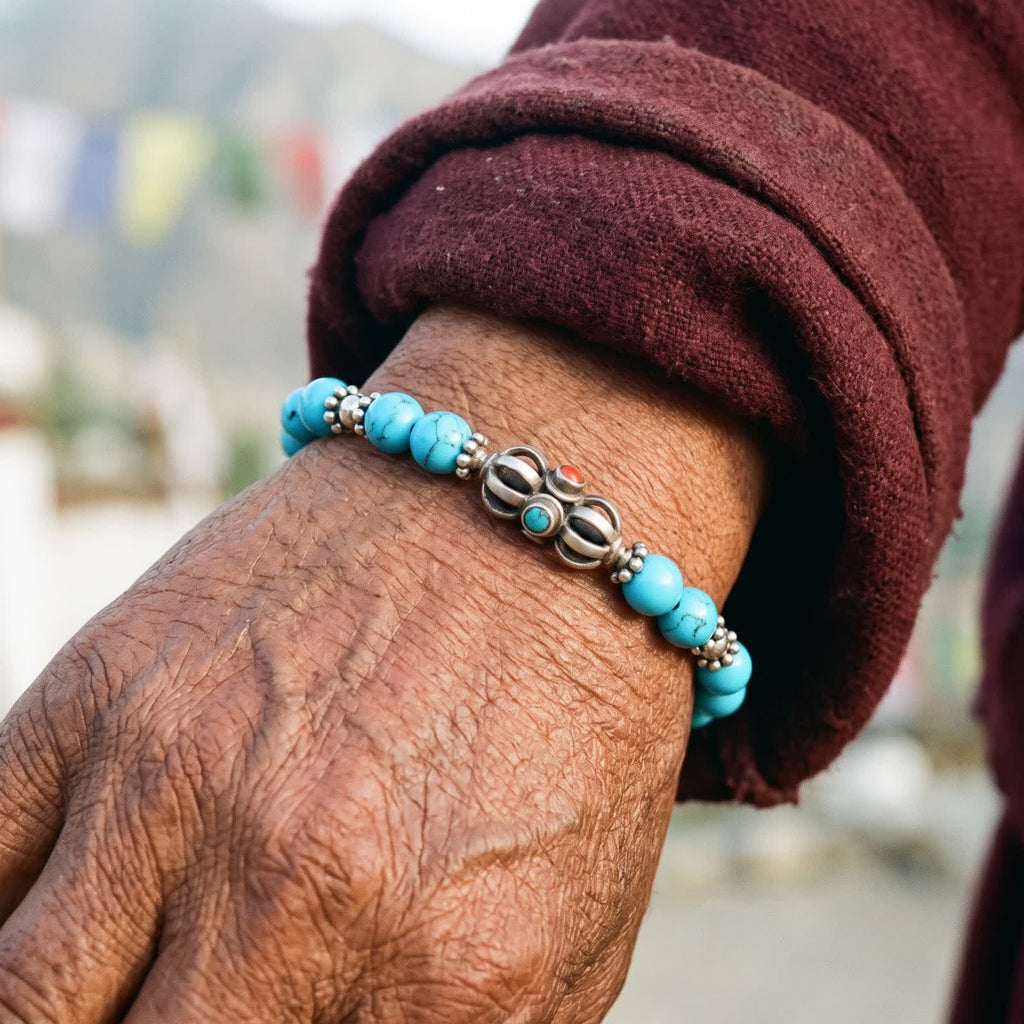 Weathered hand in a maroon wool sleeve wearing the blue turquoise bracelet with its silver vajra, prayer flags behind