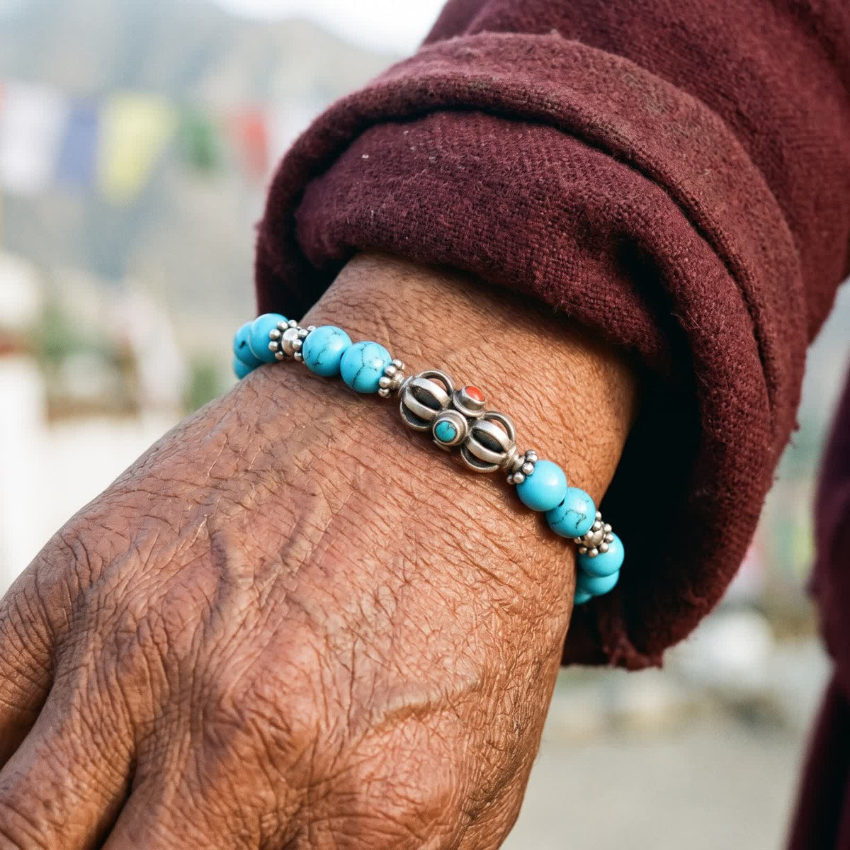 Weathered hand in a maroon wool sleeve wearing the blue turquoise bracelet with its silver vajra, prayer flags behind