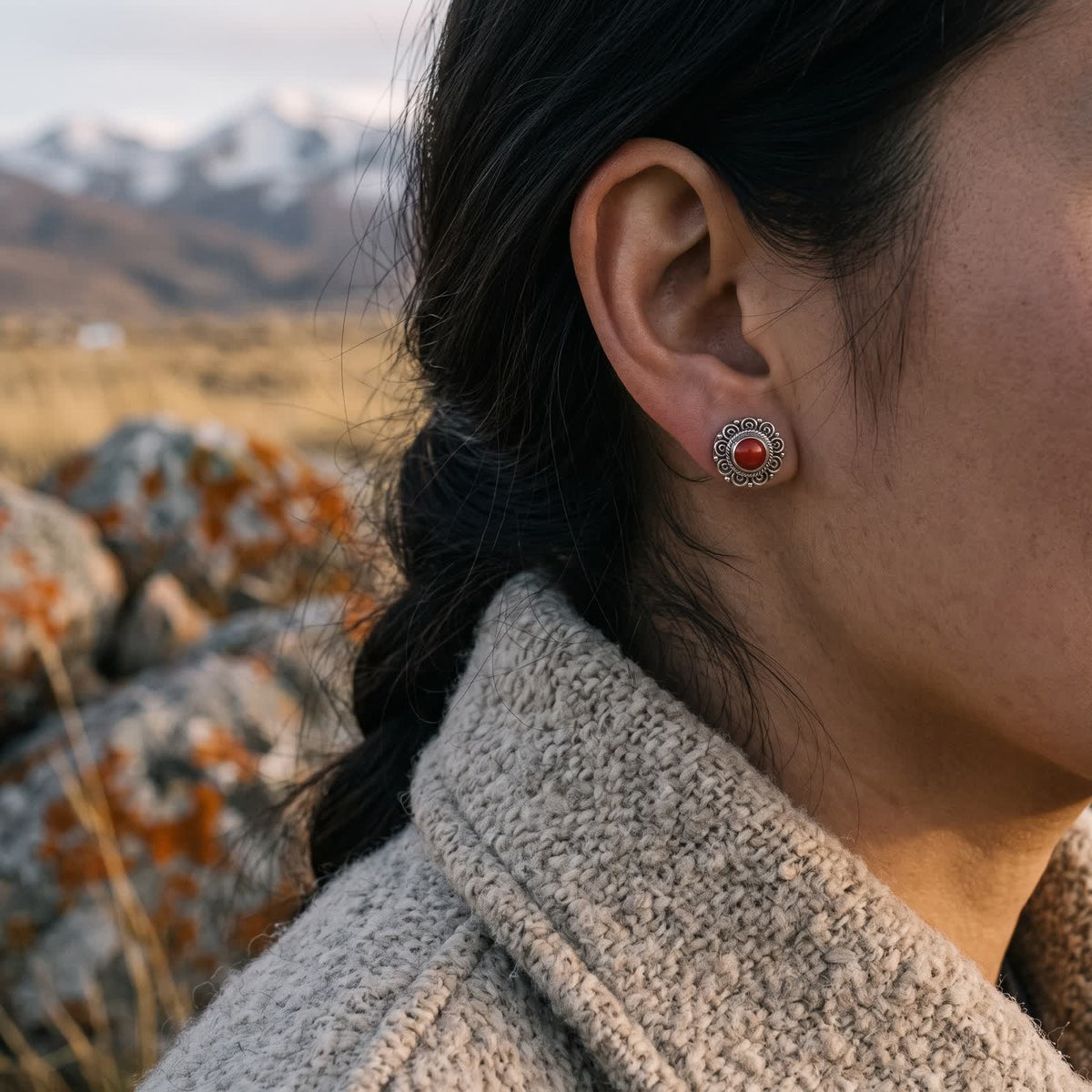 Side view of a red stone filigree stud on an ear above an oatmeal wool collar and a dark braid, snow peaks blurred behind