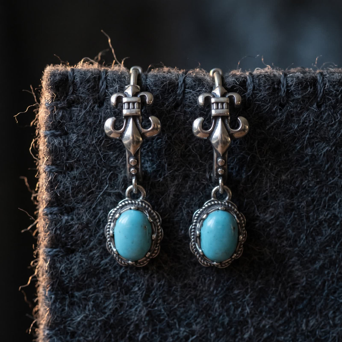 Two fleur-de-lis hoop earrings with oval turquoise drops hanging from the edge of dark charcoal wool felt