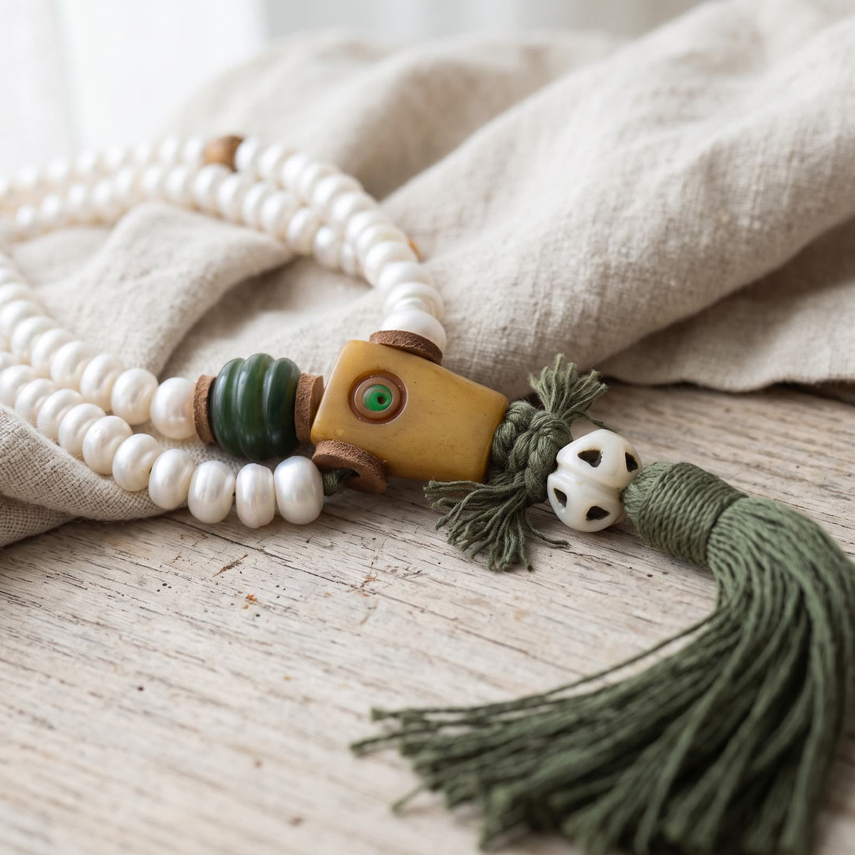 Squared guru bead with a small green inlaid dot, a green ribbed bead and leather spacers, above an openwork white bead and a dark green tassel