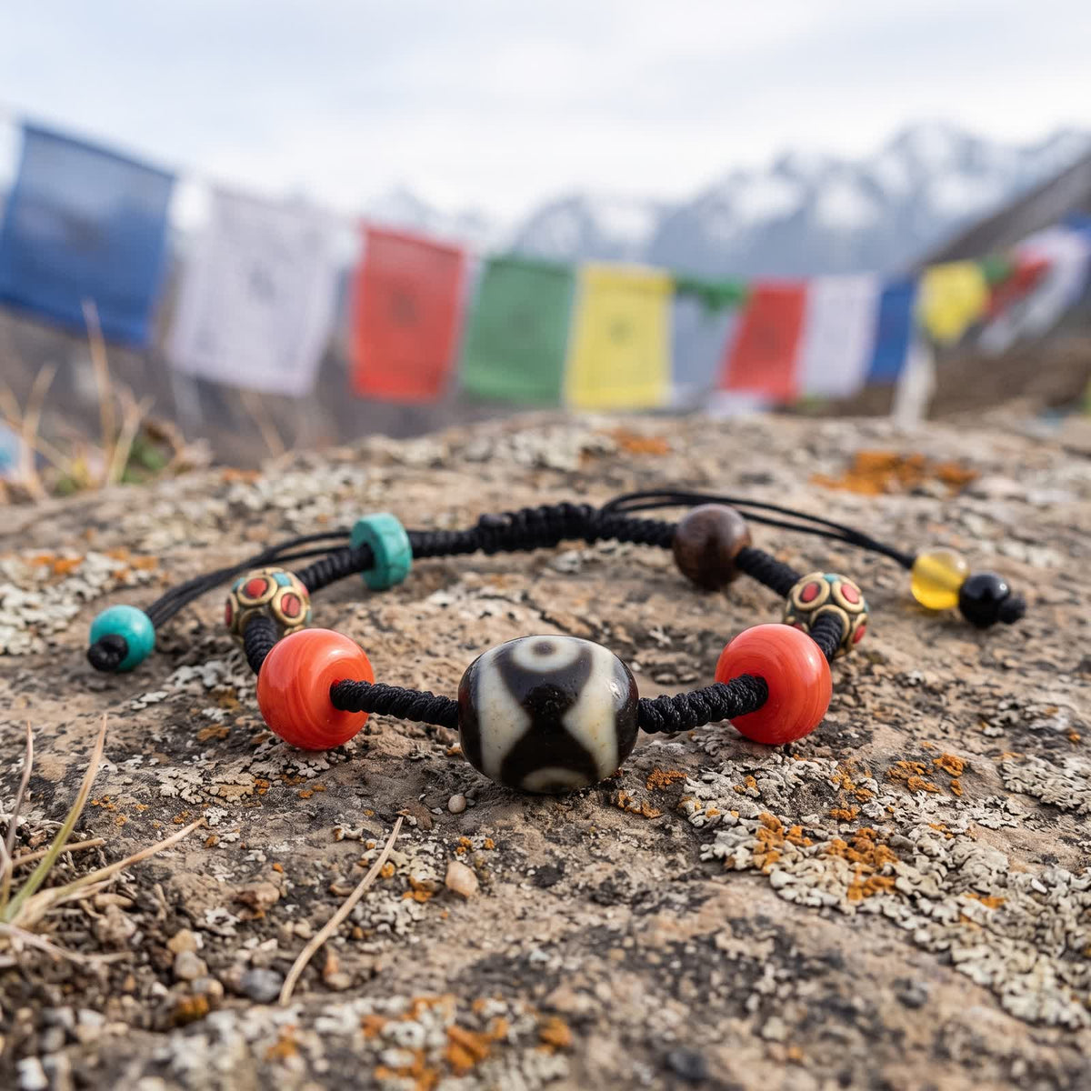 Dalo dzi cord bracelet on a weathered rock, a string of prayer flags and snow peaks blurred behind