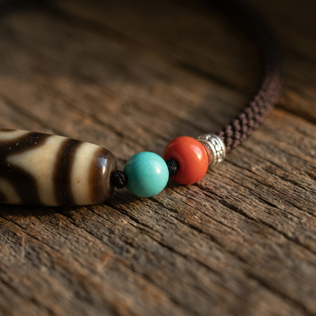 Close-up of a blue-green bead, a red bead and an embossed spacer on the braided cord beside the end of the agate dzi bead