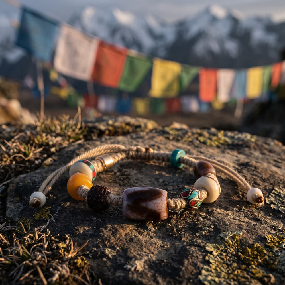 Tiger-tooth dzi cord bracelet on a weathered rock in warm evening light, prayer flags and snow peaks blurred behind