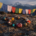 Three-eyed red dzi cord bracelet on a lichen-covered rock in warm evening light, prayer flags and snow peaks blurred behind