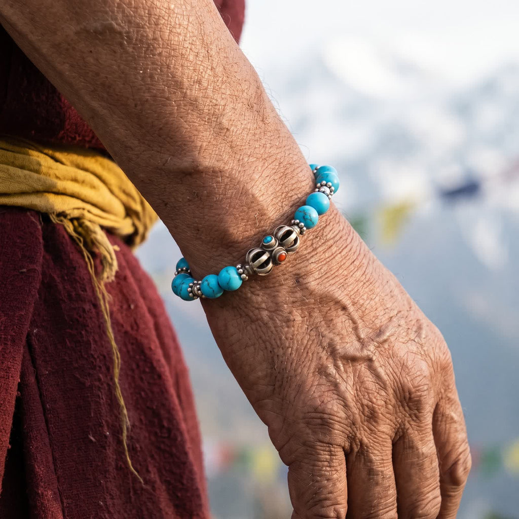 Older wrist at the side of a maroon robe and yellow sash wearing the blue turquoise and silver vajra bracelet, snowy peaks behind