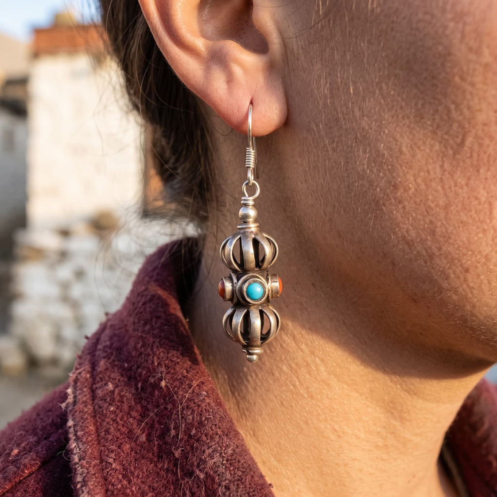 Vajra drop earring on a U-shaped ear wire, worn on a woman's ear above a maroon wool collar, late-afternoon light