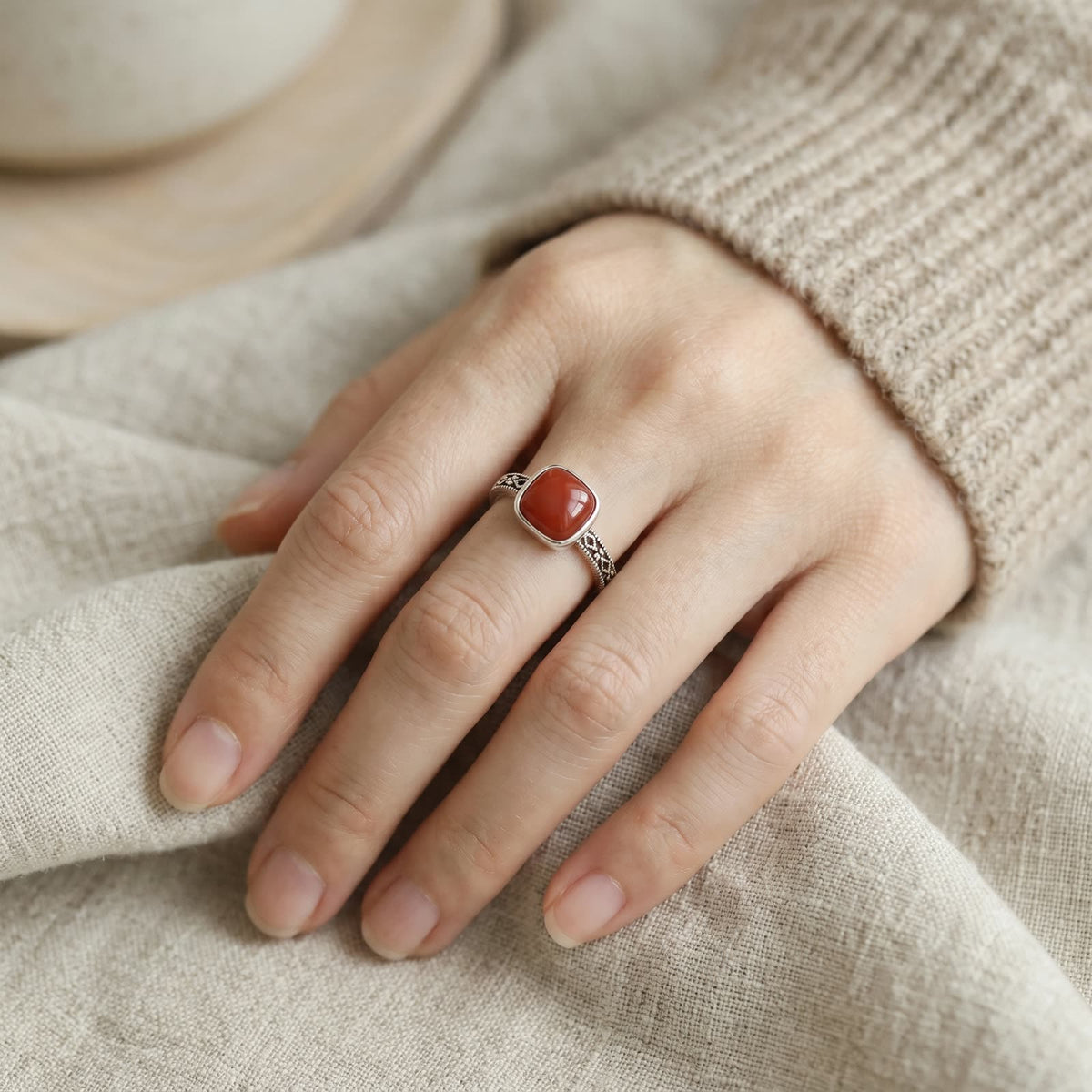 Openwork ring with a square red stone worn on the middle finger of a relaxed hand resting on linen