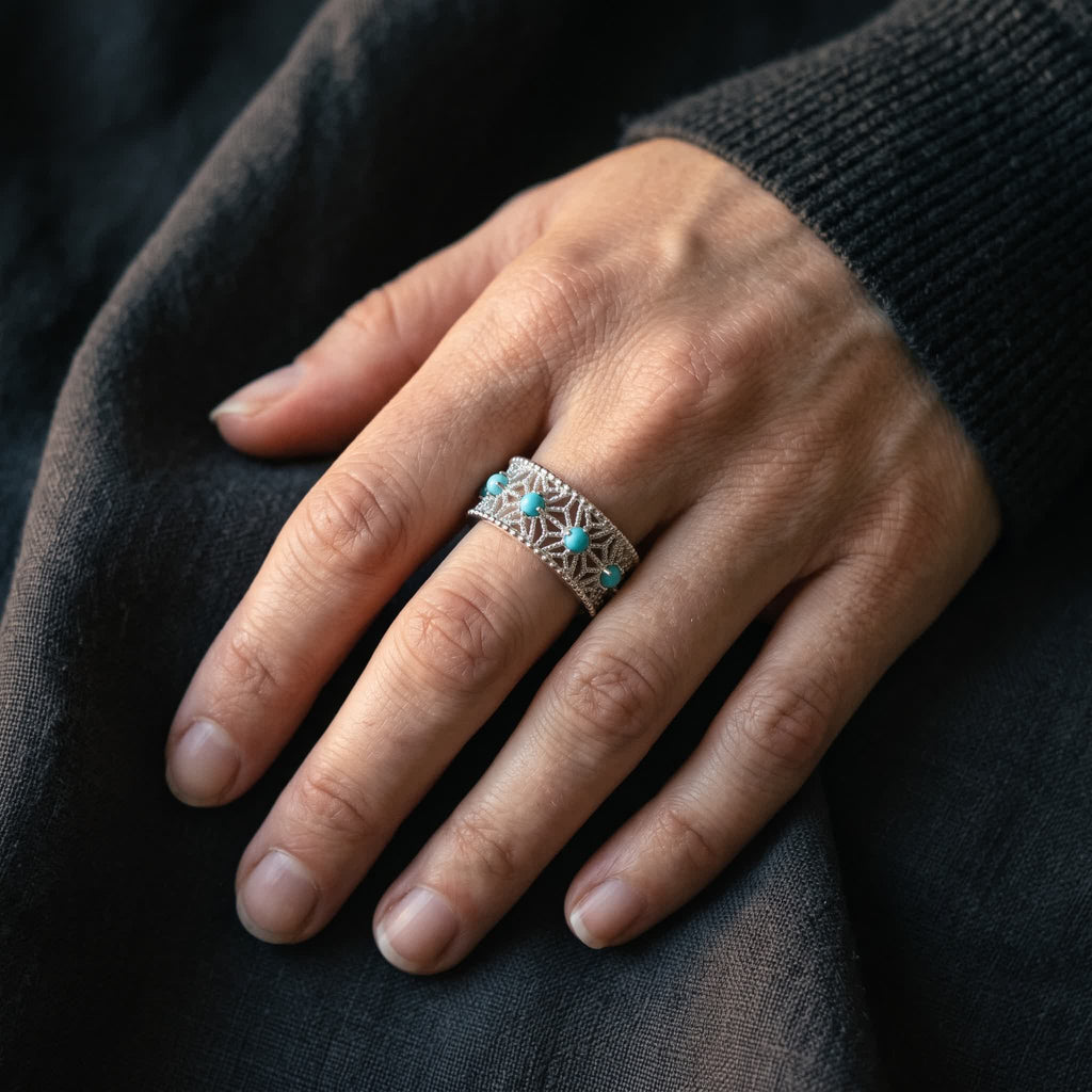 Wide openwork sterling silver ring with blue-green stones worn on the middle finger of a relaxed hand on dark linen