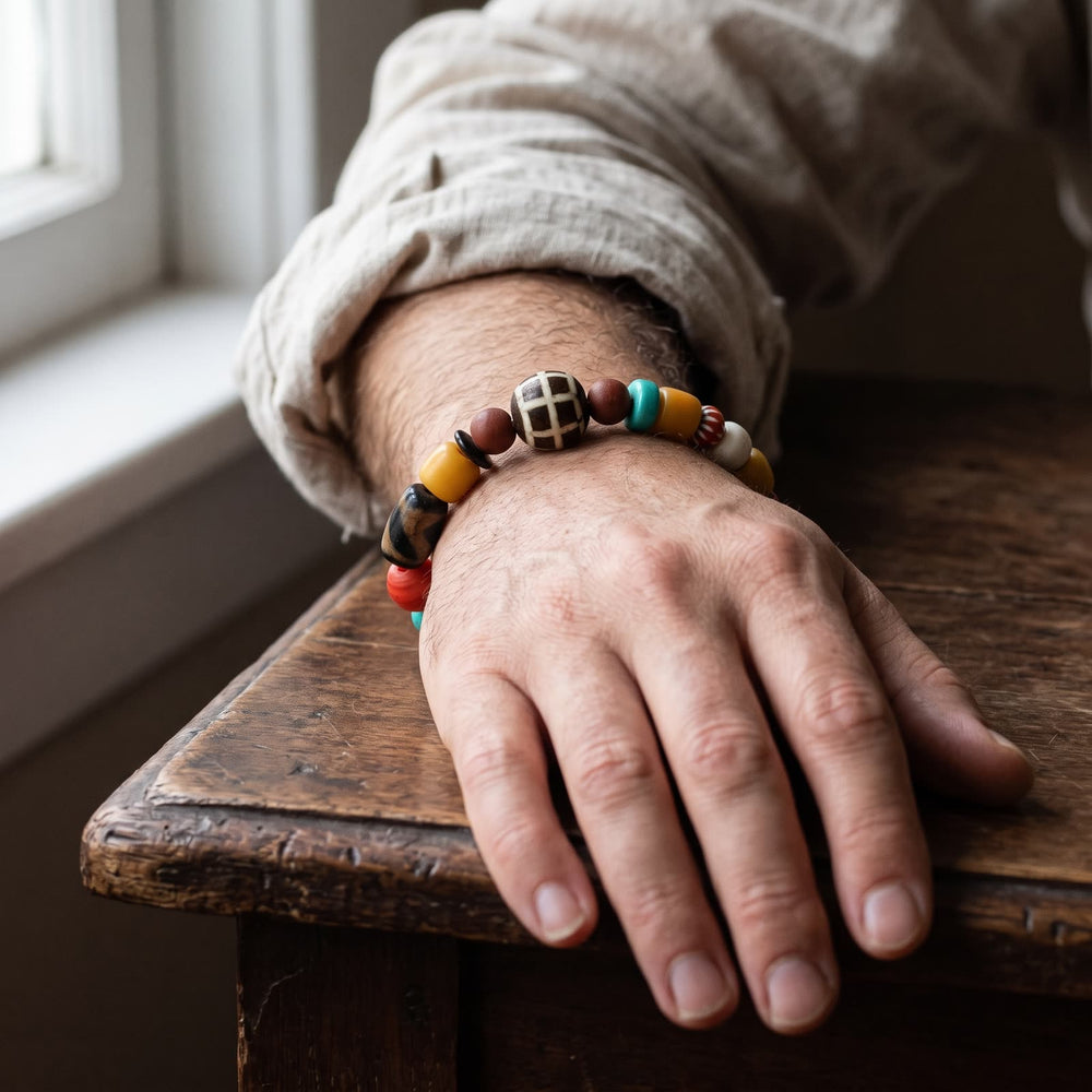Multi-bead bracelet on a man's wrist showing the grid-patterned yak bone bead, linen sleeve rolled back, hand resting on a wooden table