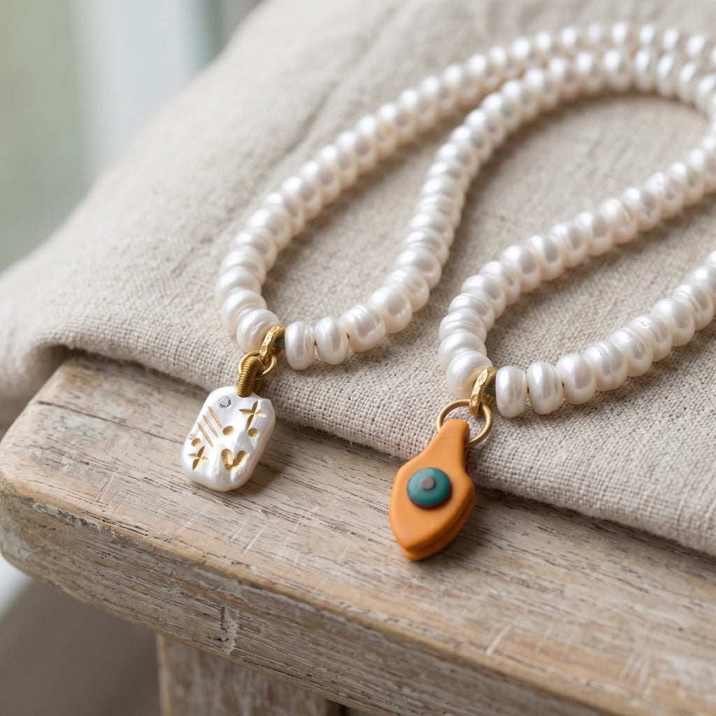 Golden engraved white charm and an orange drop charm with a teal dot, hanging from pearl-white beads on undyed linen