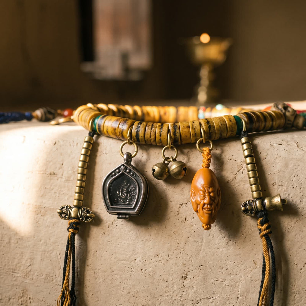 Hanging charms on the bone disc mala: two golden bead strings with small metal finials, a gau box pendant, two small golden bells and an orange-brown carved face bead