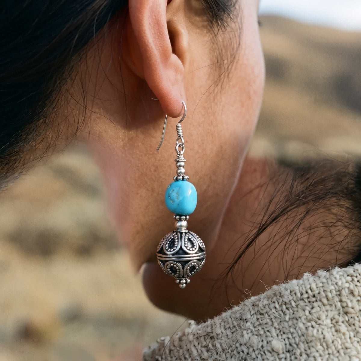 Turquoise and filigree ball drop earring worn on a woman's ear, hair tucked back, warm hills blurred behind