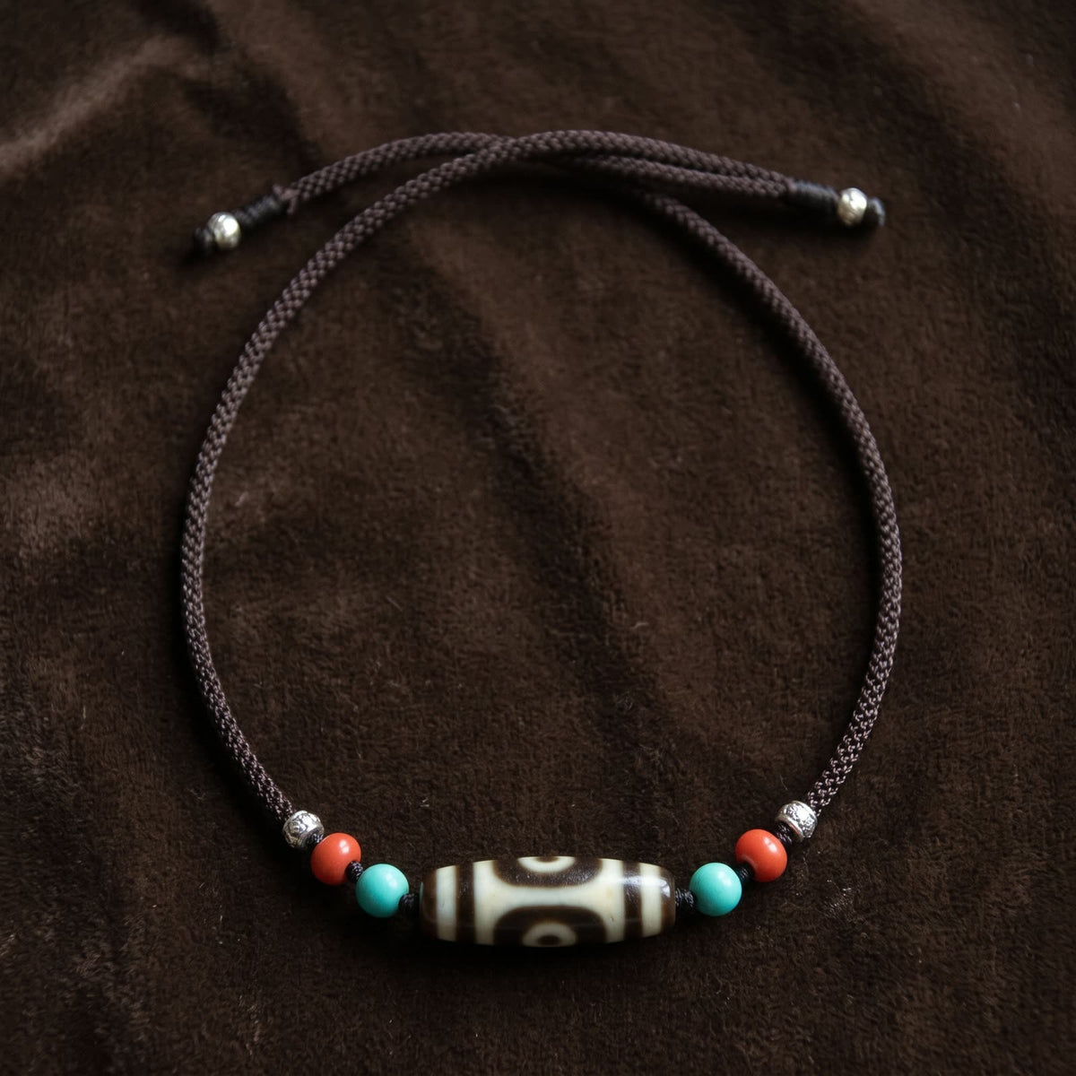 Full-length flat lay of the two-eyed agate dzi necklace on dark brown velvet, showing the braided cord and the two adjustable sliding knots with beads