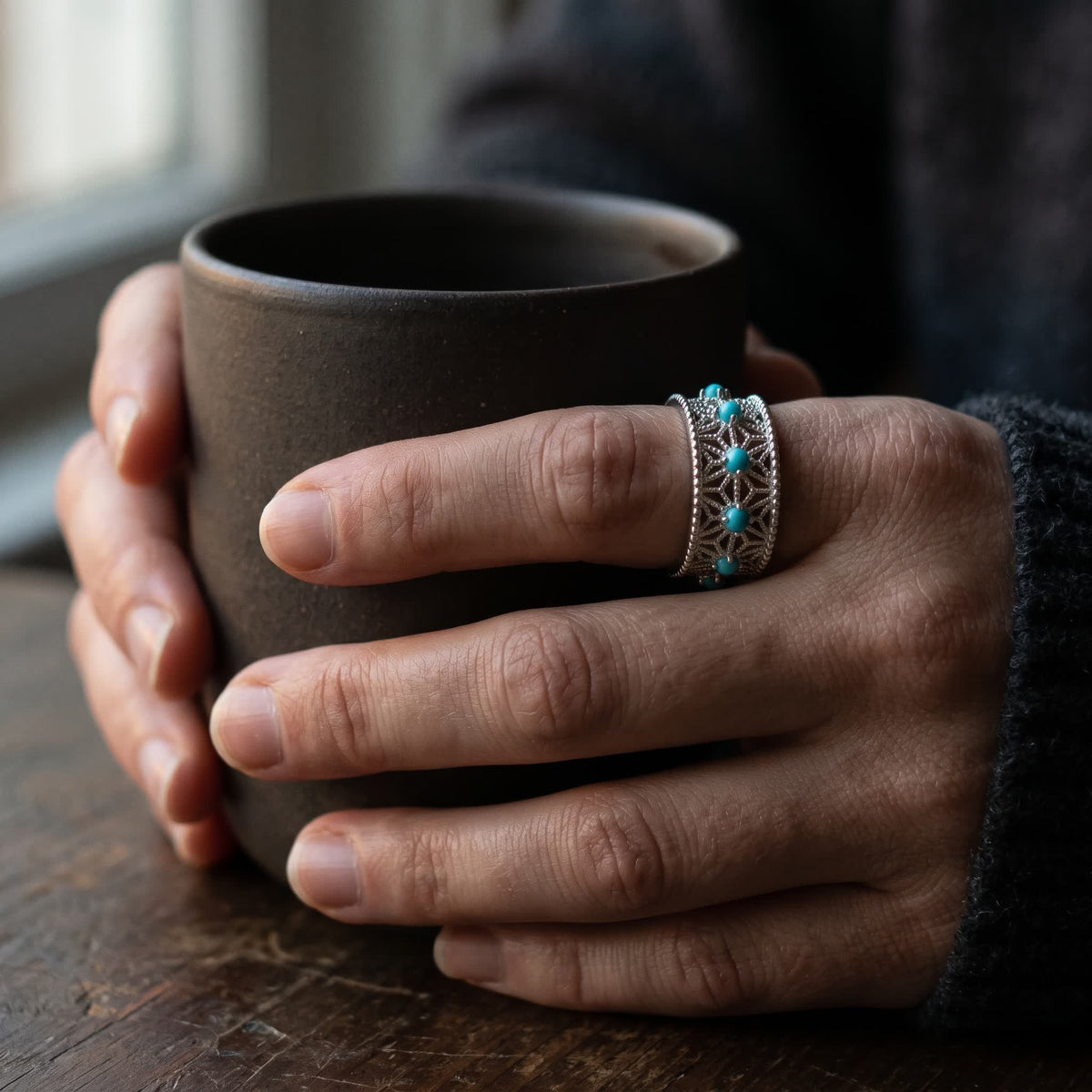 Hands holding a dark stoneware cup, the openwork silver ring with a row of blue-green stones on the index finger