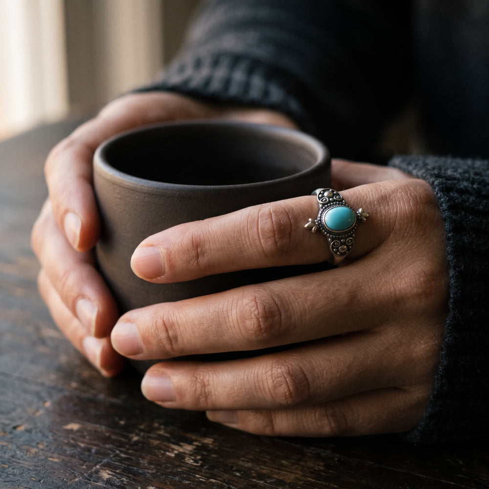 Hands holding a dark stoneware cup, an oval turquoise ring with darkened scrollwork on the index finger