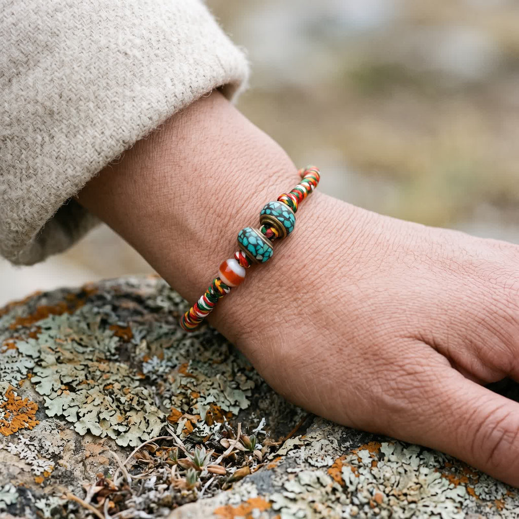Five-color cord bracelet worn on a wrist below an oatmeal wool sleeve, its banded agate and turquoise-chip inlay copper beads on top, hand on a lichen-covered rock