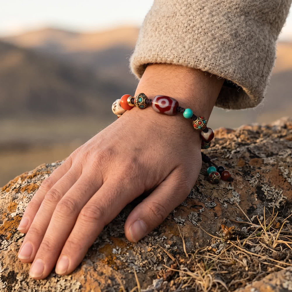 Three-eyed red dzi cord bracelet worn on a wrist below an oatmeal wool sleeve, hand resting on a lichen-covered rock, cord tails with small beads hanging down