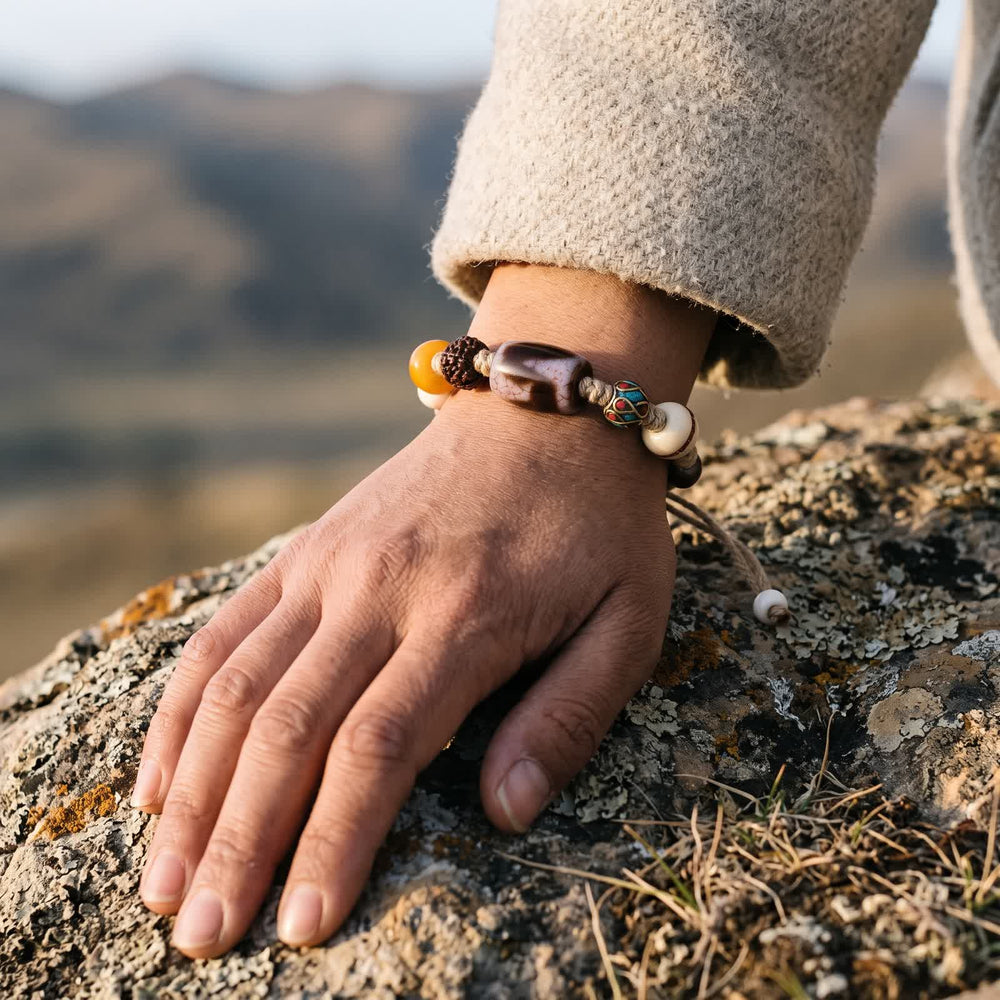 Tiger-tooth dzi cord bracelet worn on a wrist below an oatmeal wool sleeve, hand resting on a lichen-covered rock in soft sunlight
