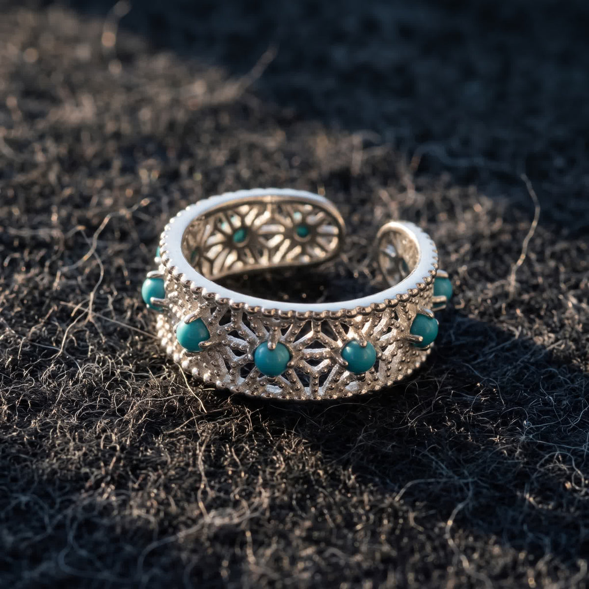 Adjustable openwork silver ring with blue-green stones lying on dark charcoal wool felt in warm side light