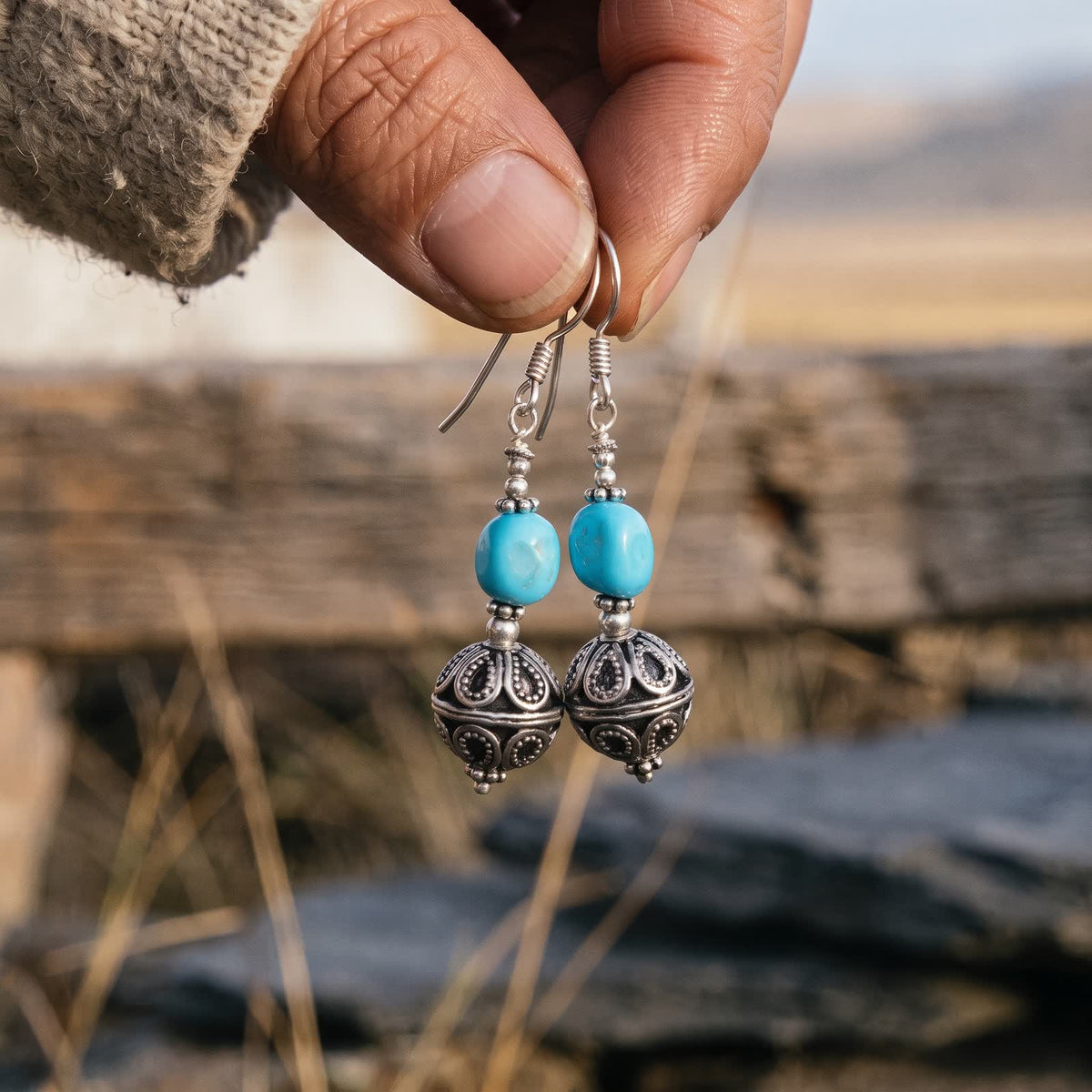 Pair of turquoise filigree ball drop earrings hanging from a woman's fingertips in warm evening light