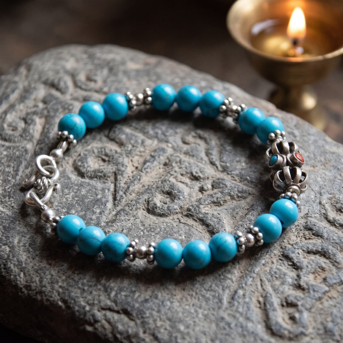 Blue turquoise bracelet with a silver vajra resting on a carved grey stone beside a glowing butter lamp