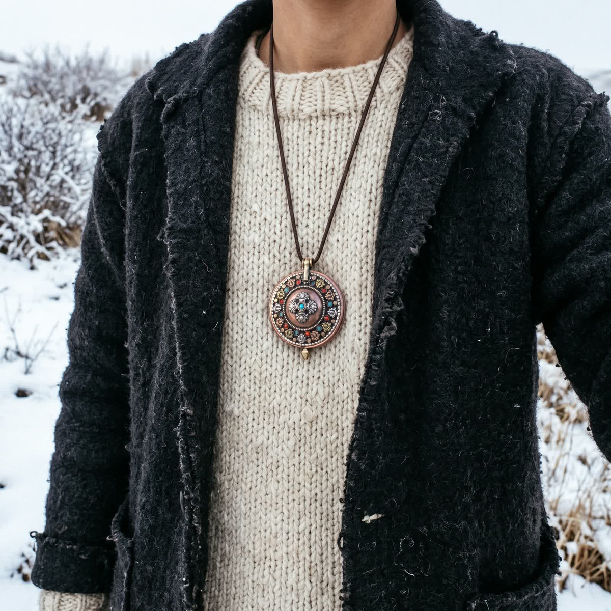 Copper gau box pendant worn on a thin brown cord over a cream wool sweater and dark wool coat, snowy field behind