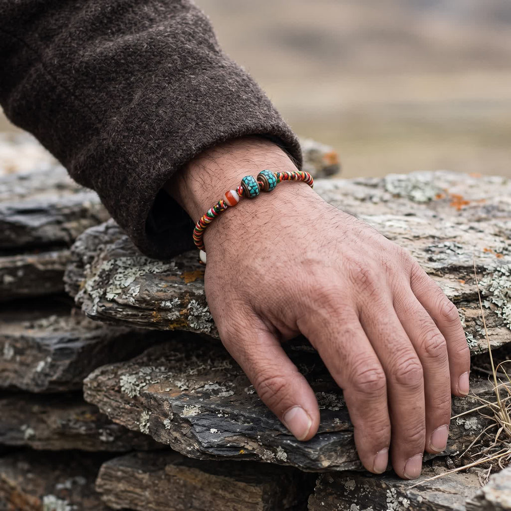 Five-color cord bracelet worn on a man's wrist below a brown wool sleeve, hand resting on stacked slate
