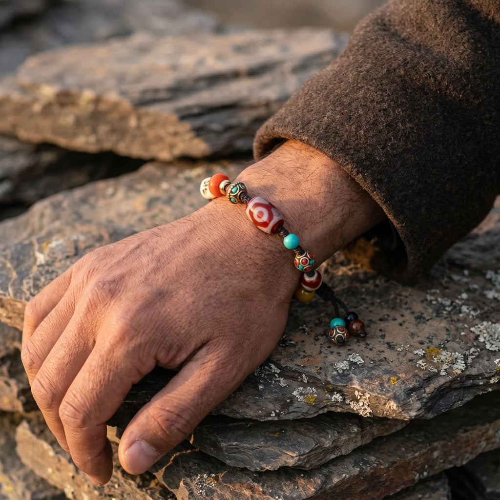 Three-eyed red dzi cord bracelet on a man's wrist below a brown wool sleeve, hand resting on stacked slate in warm light