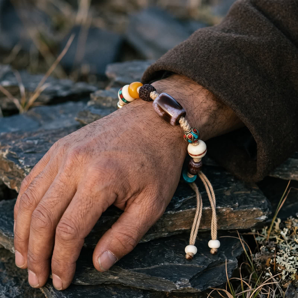 Tiger-tooth dzi cord bracelet on a man's wrist below a brown wool sleeve, two cord tails with small white end beads hanging over dark slate