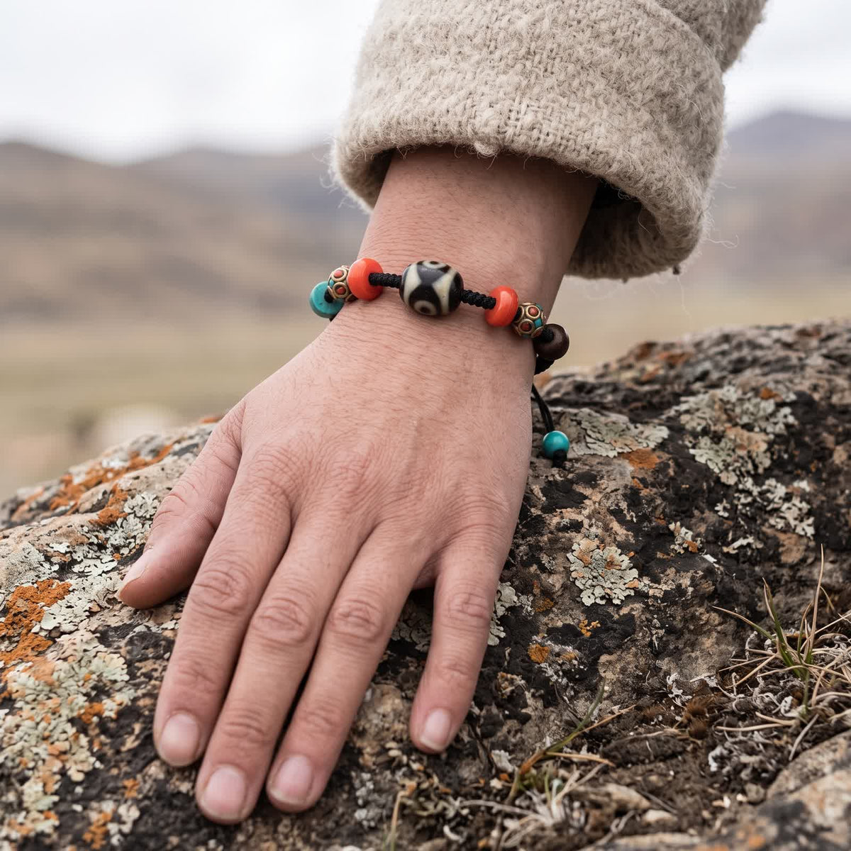 Dalo dzi cord bracelet worn on a wrist below an oatmeal wool sleeve, hand resting on a lichen-covered rock