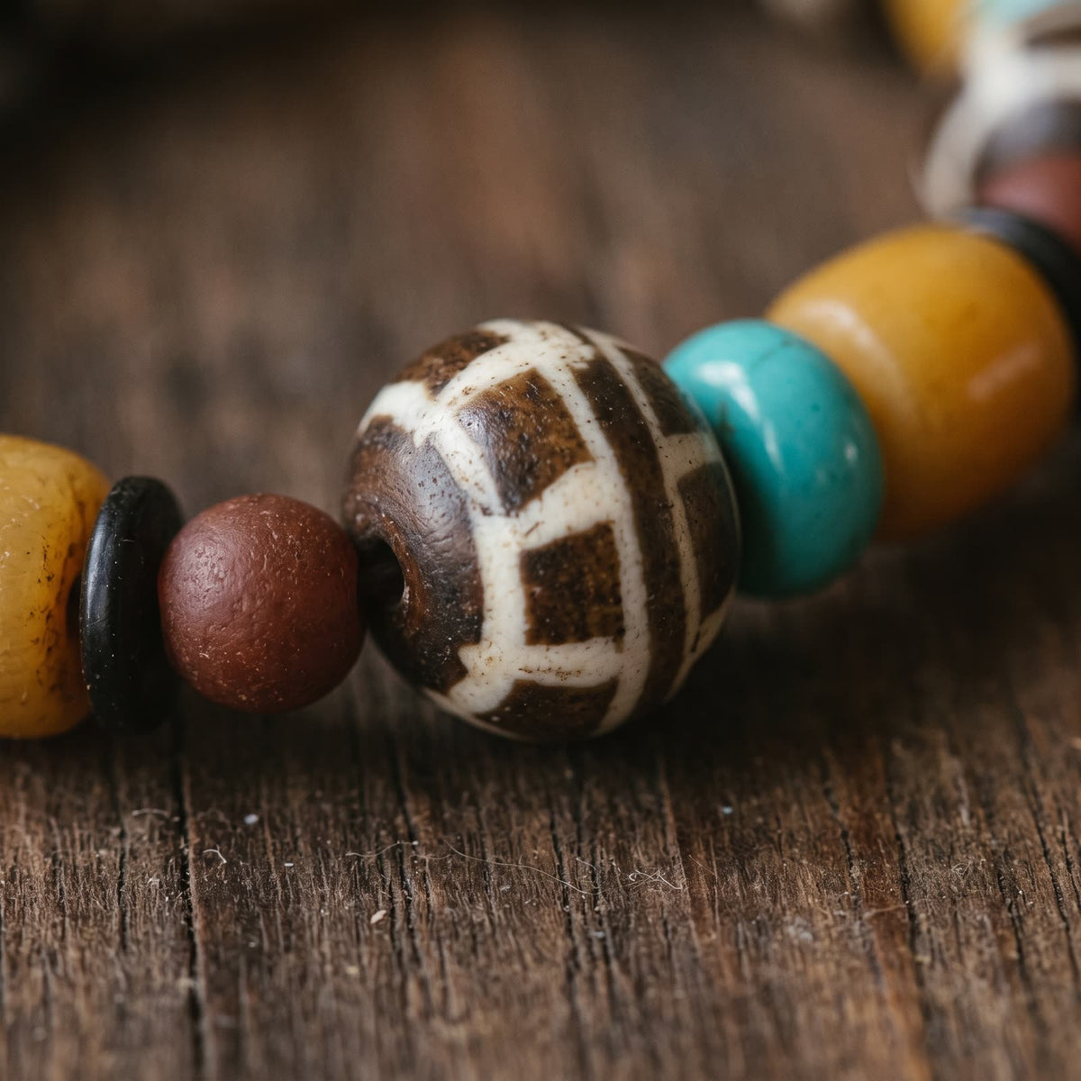 Close-up of the round yak bone bead with a white grid on dark brown, beside turquoise and yellow ox bone beads on weathered wood