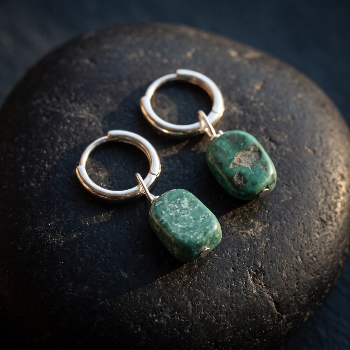 Two green stone hoop earrings with different markings resting on a black river stone
