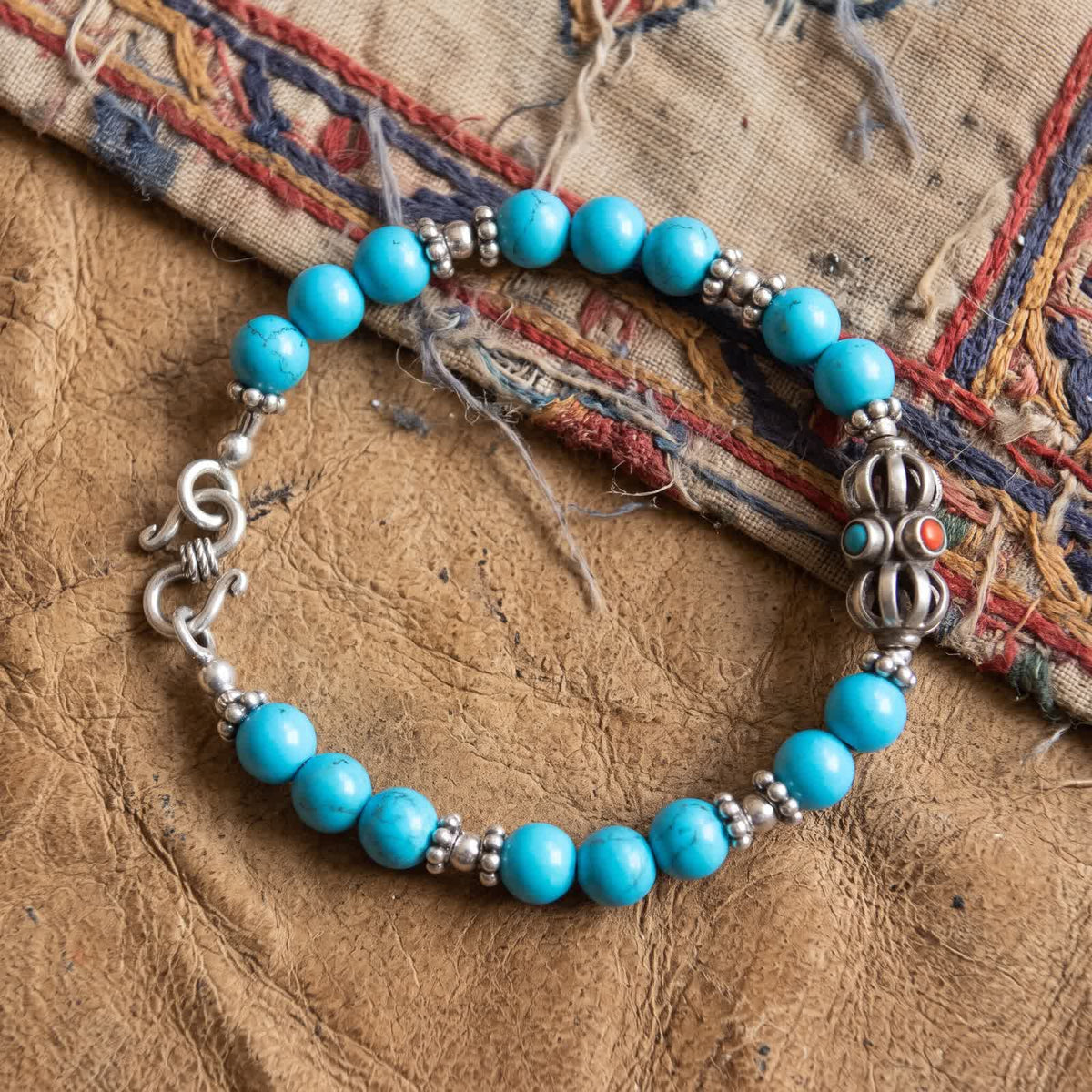 Top-down view of the blue turquoise bracelet with small silver spacers and a silver vajra on worn leather and embroidered cloth