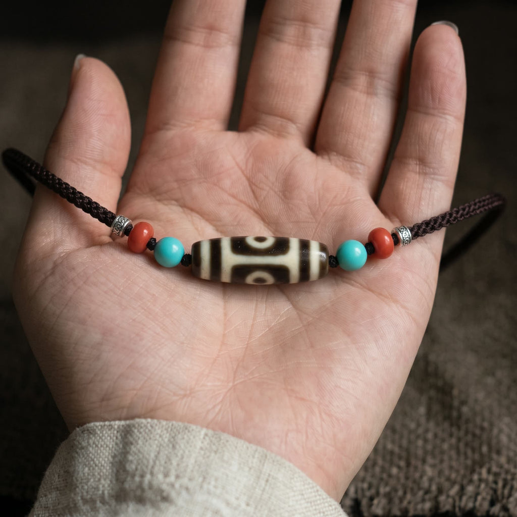 Open palm holding the agate dzi bead with its blue-green and red beads to show its size, braided cord falling from the hand