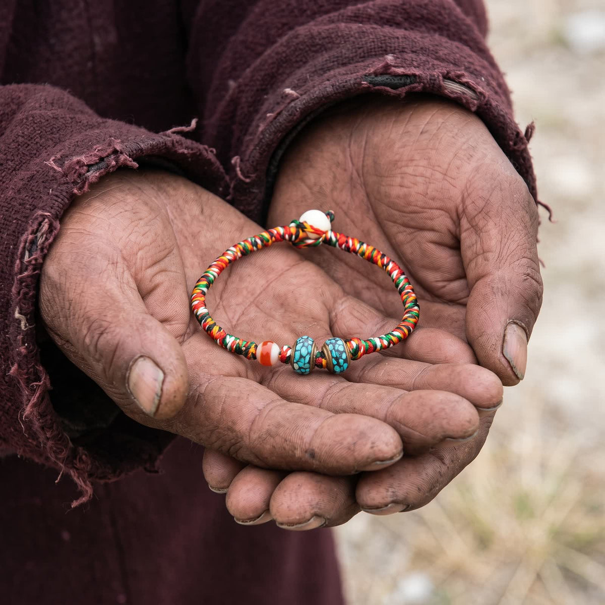 Weathered hands in a worn maroon wool robe holding the five-color cord bracelet in open palms