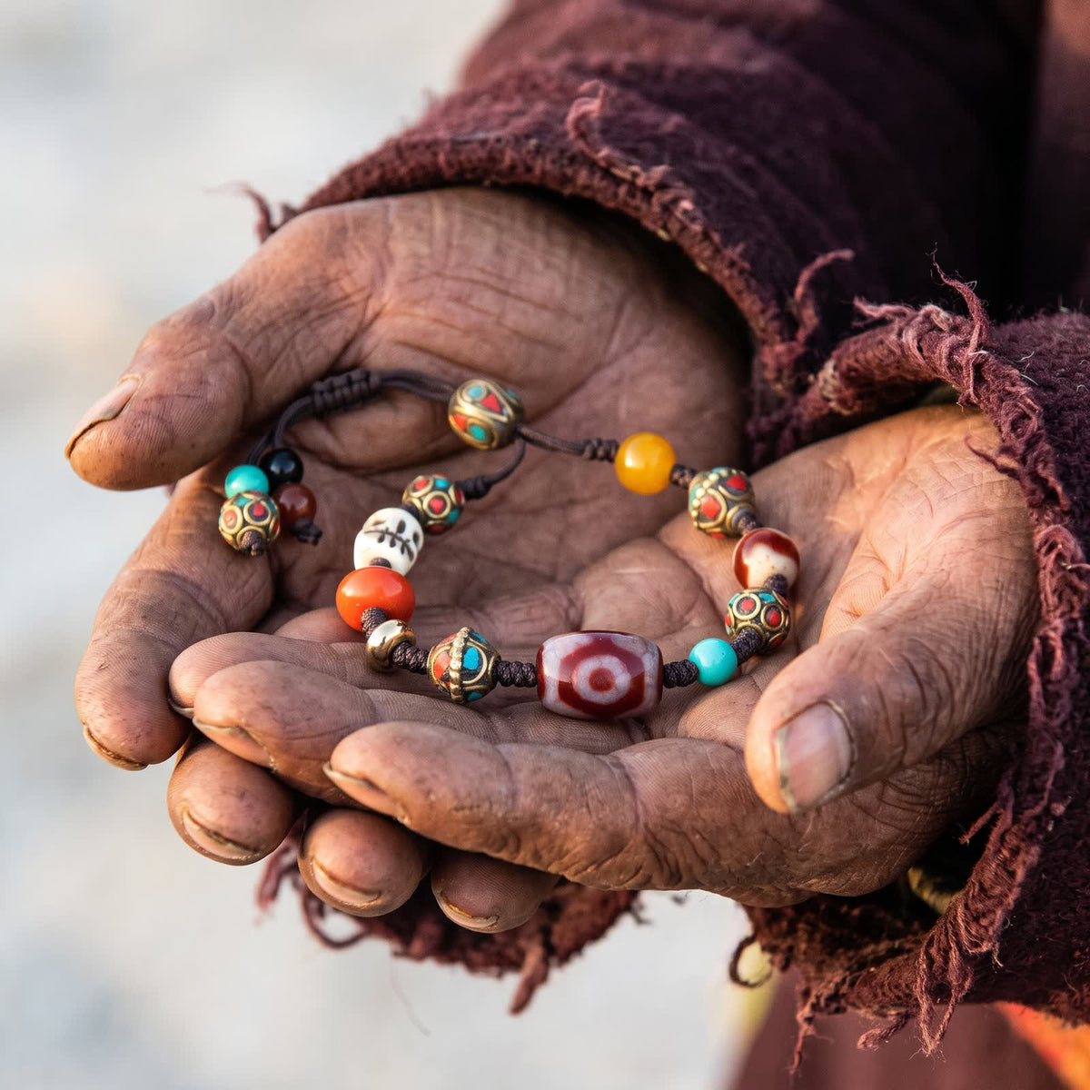 Weathered hands in a worn maroon wool robe holding the three-eyed red dzi cord bracelet in cupped palms