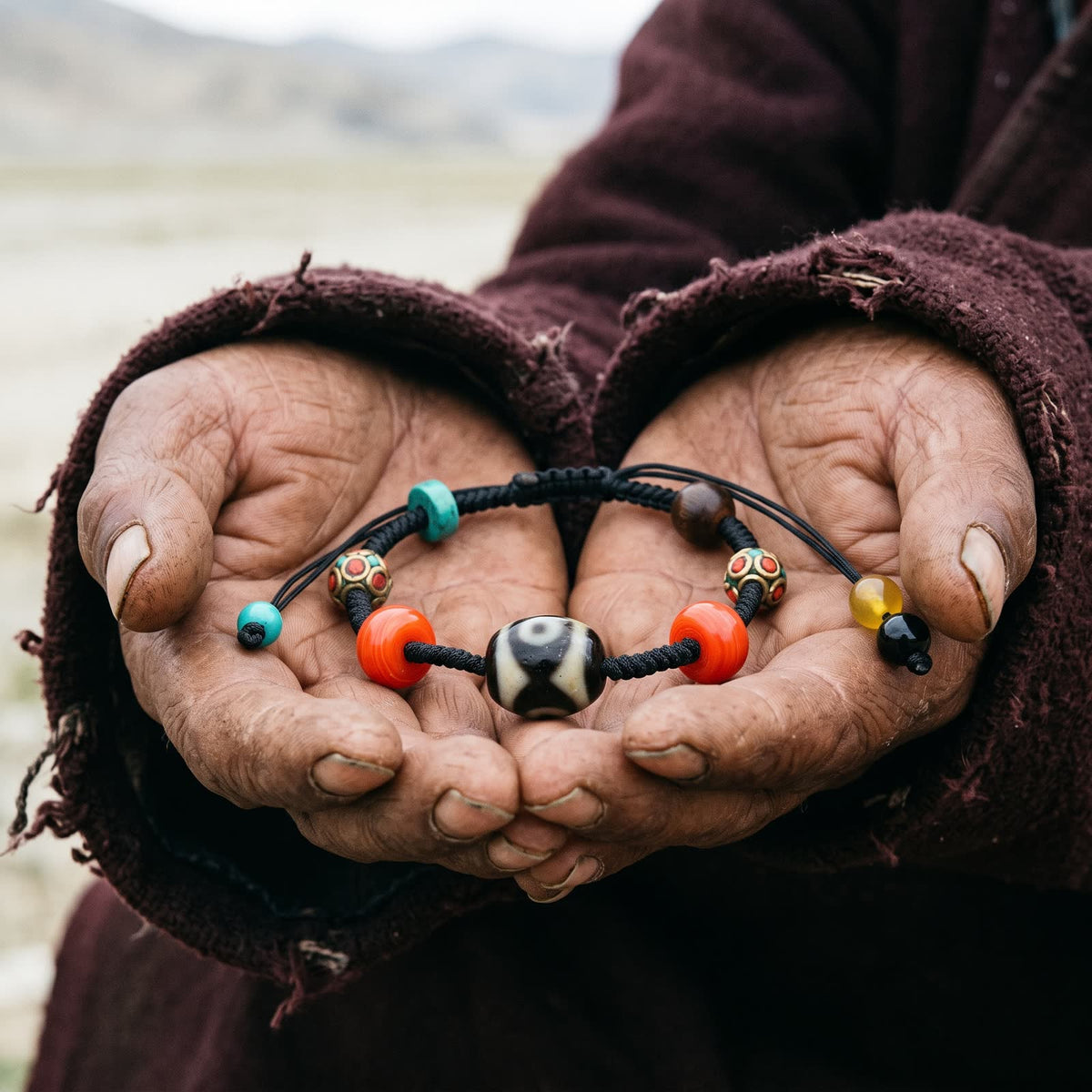 Weathered hands in a worn maroon wool robe holding the Dalo dzi cord bracelet in open palms outdoors