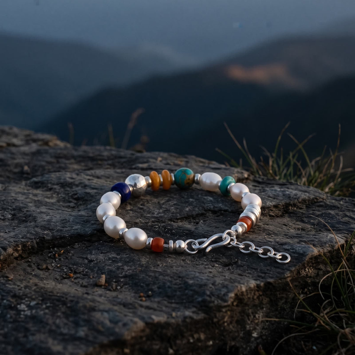 Pearl bracelet lying on a dark rock ledge at dusk, layered blue mountain ridges softly out of focus behind