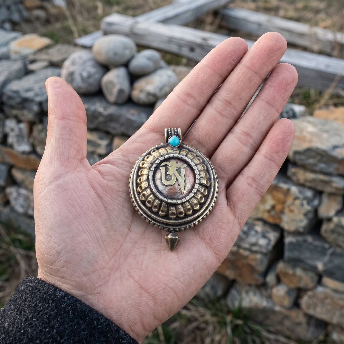 Om gau box pendant resting in an open palm for scale, about 4.4 cm across, with a stone wall behind