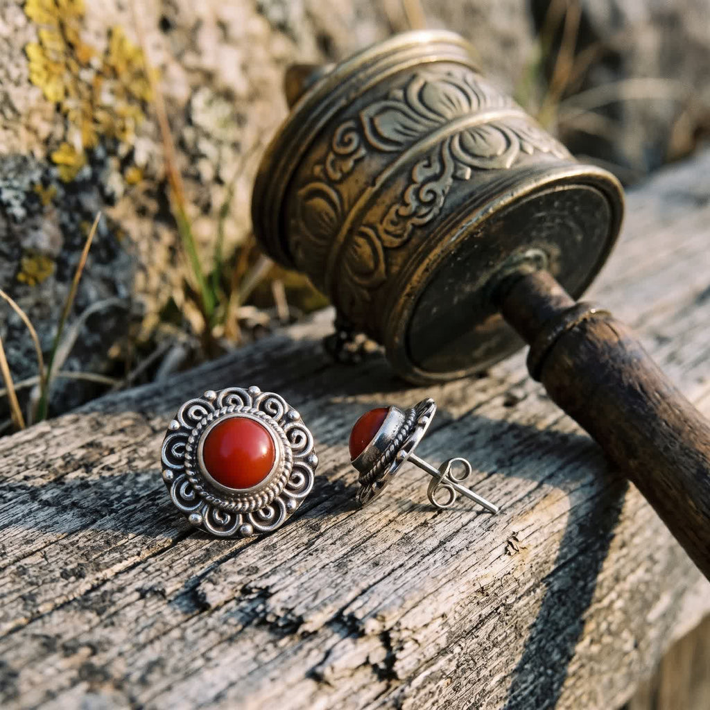 Red stone filigree stud earrings on weathered wood beside a brass hand prayer wheel, one turned to show its post