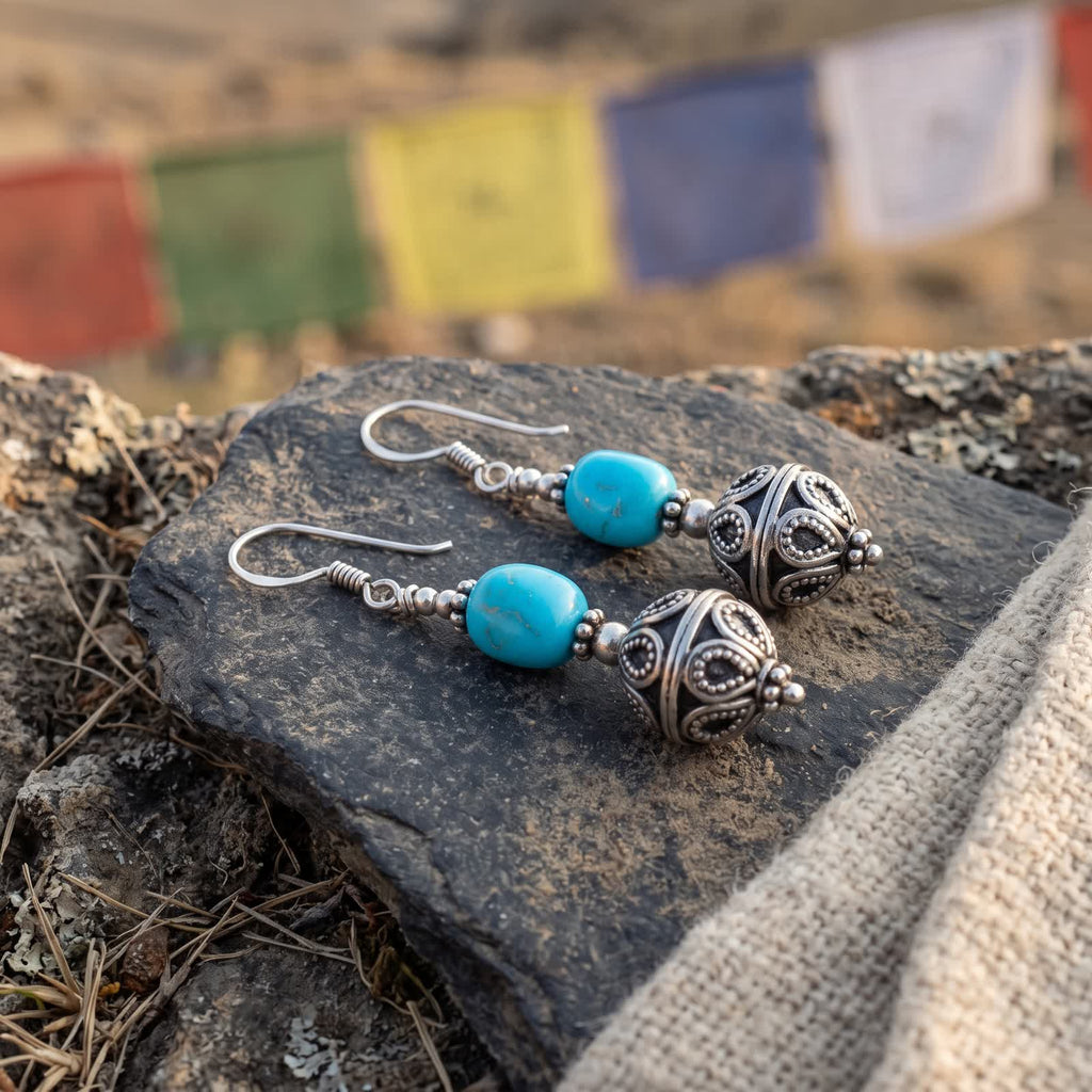 Turquoise and silvery filigree ball earrings on a slate stone beside undyed wool, prayer flags behind