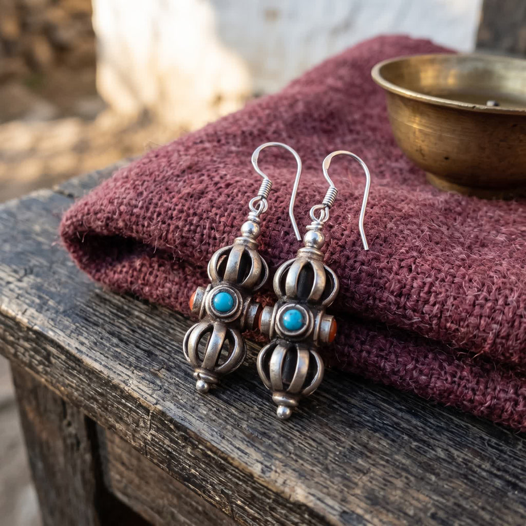 Pair of vajra drop earrings with red and blue stones on folded maroon wool beside a small brass bowl