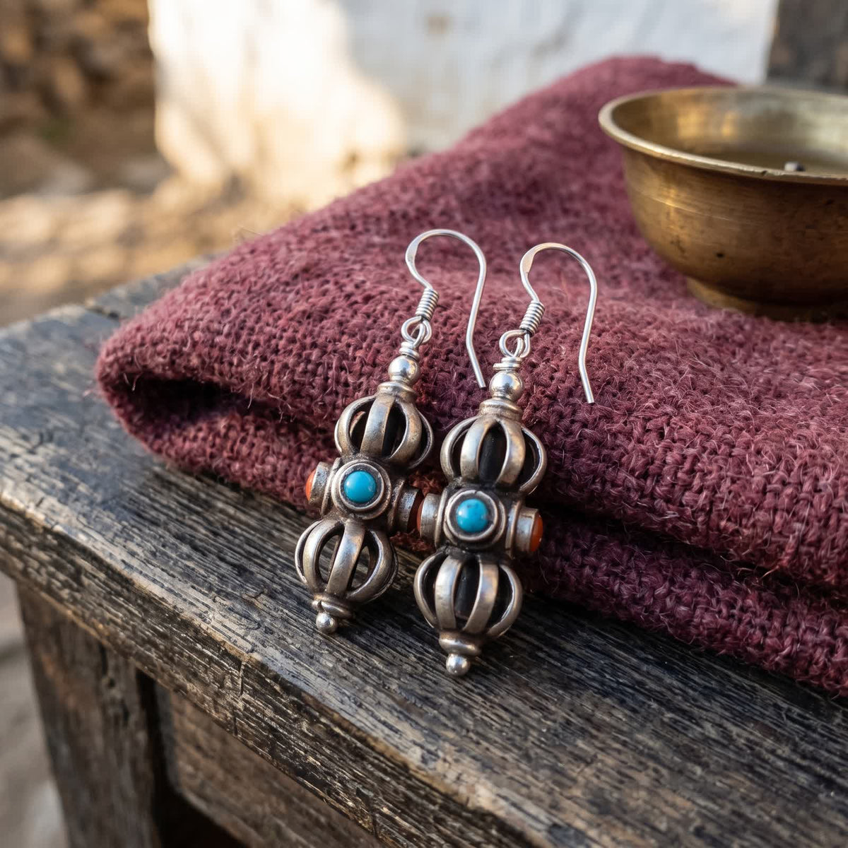 Pair of vajra drop earrings with red and blue stones on folded maroon wool beside a small brass bowl