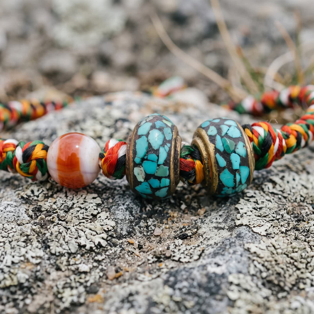 Close-up of the red-and-white banded agate bead and two copper beads inlaid with turquoise chips on red, yellow, green, white and black braided cord