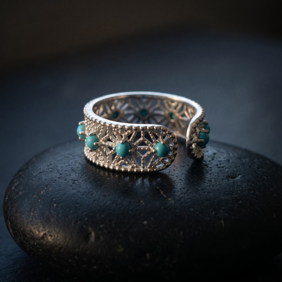 Openwork lace ring in 925 sterling silver with blue-green stones resting on a black river stone