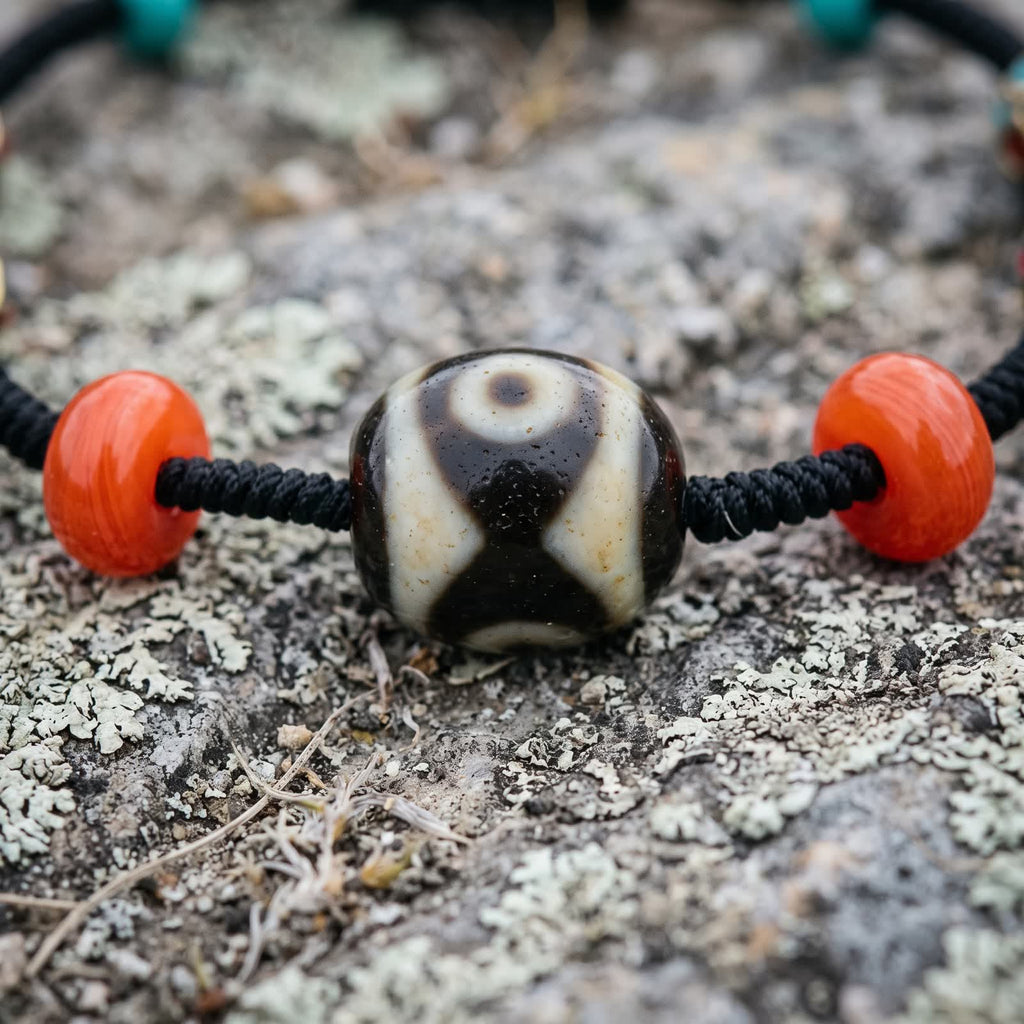 Close-up of the Dalo dzi bead, a cream eye above dark bands crossing into an X, between red glass beads on black braided cord