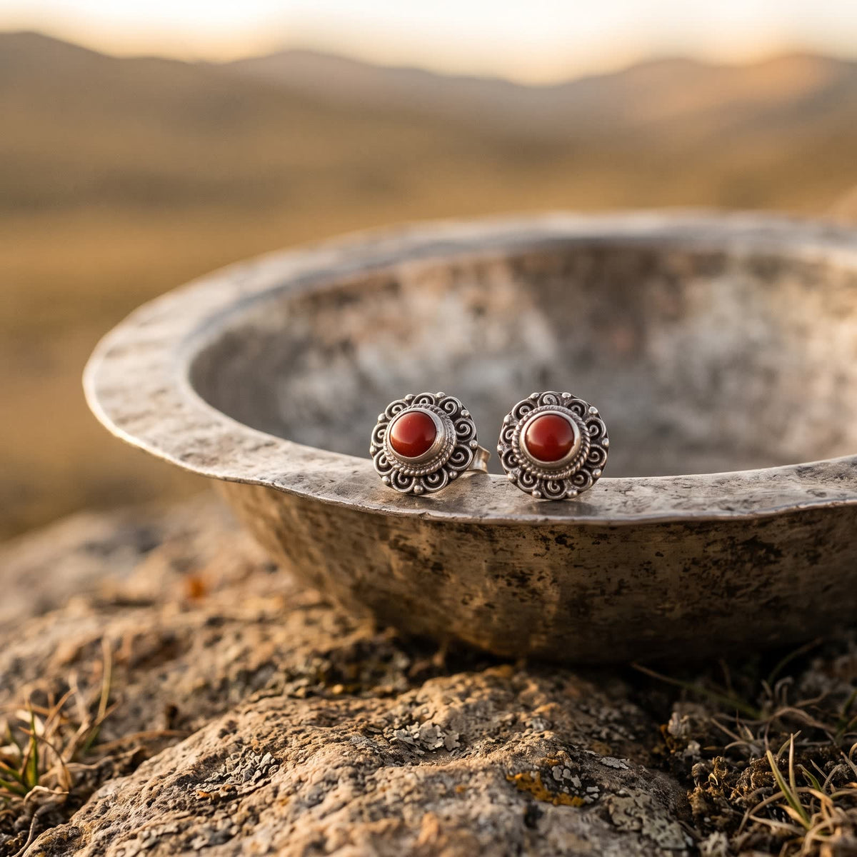 Pair of red stone filigree stud earrings resting on the rim of a hammered metal bowl, hills blurred behind in warm light