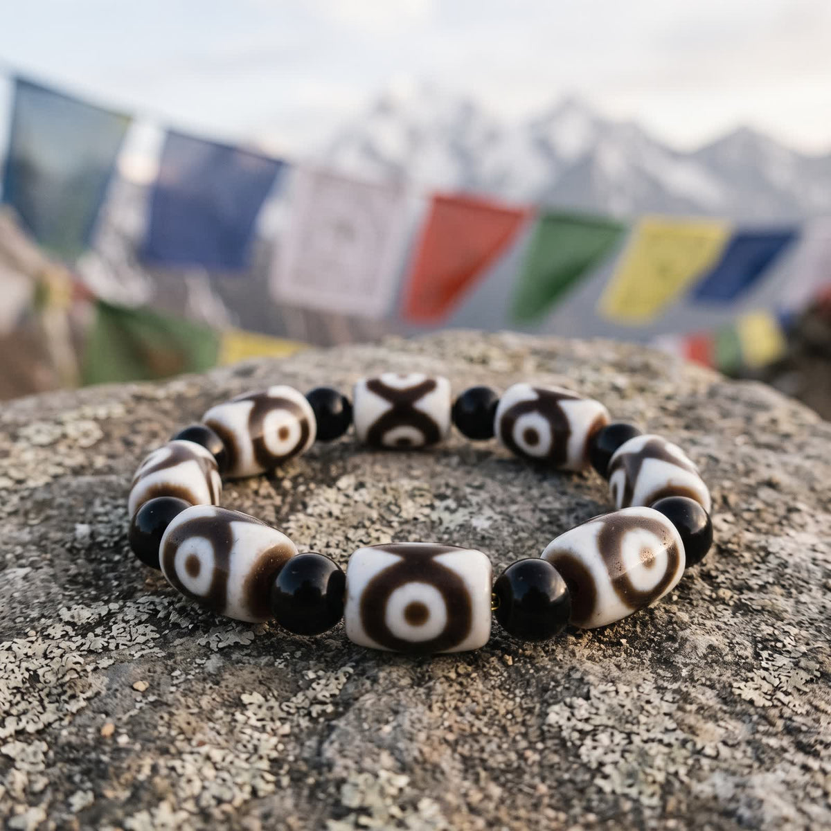 Agate dzi and black agate bead bracelet on a lichen-covered stone with prayer flags and snow peaks blurred behind